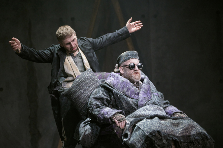 A man in a suit bowing with his arms outstretched in front of a man with dark glasses in a chair