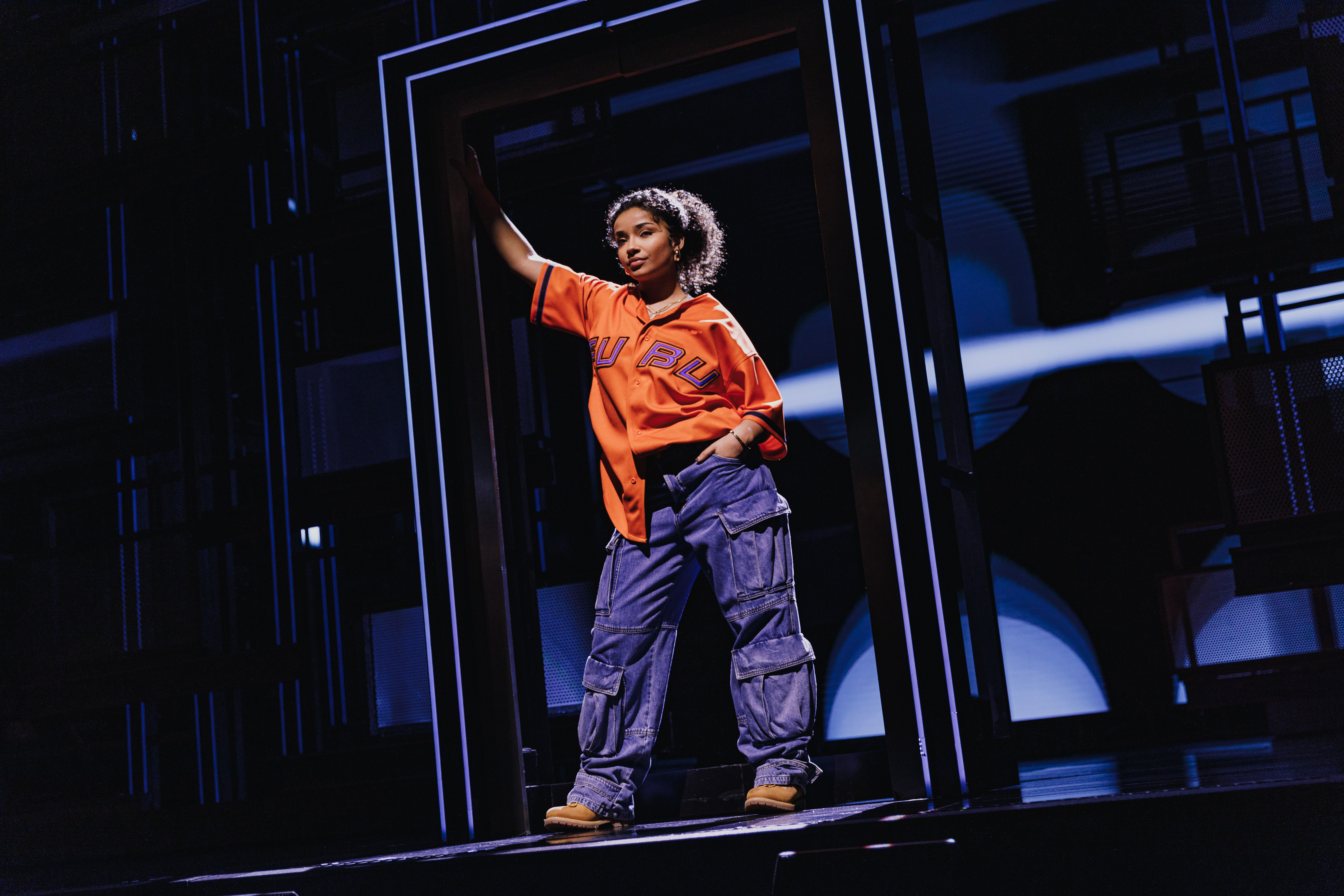 A young woman in an orange top and jeans standing in a doorway looking sassy