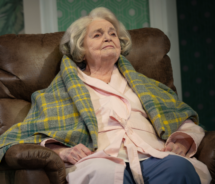 An older woman in a green plaid blanket sitting on a recliner looking pensive 