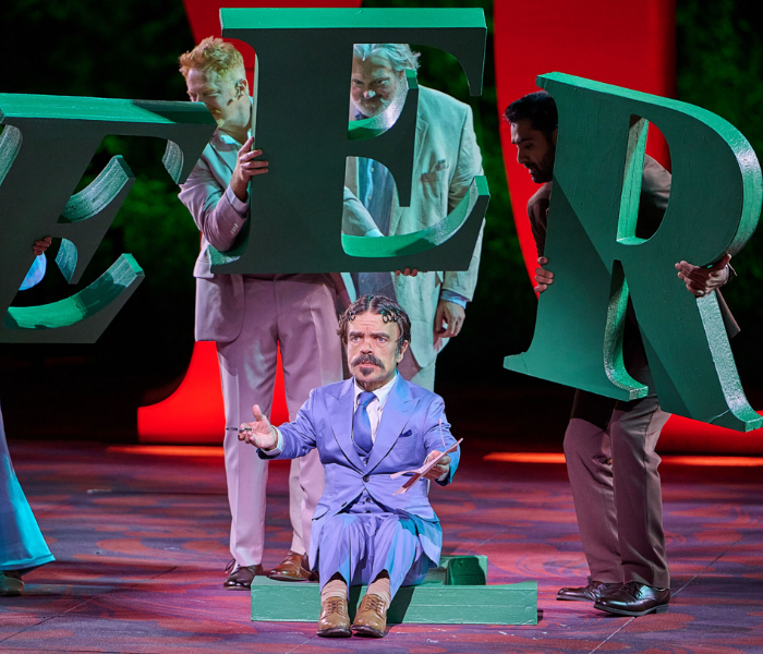Peter Dinklage in a purple suit sitting on a stage talking with large green letters behind him