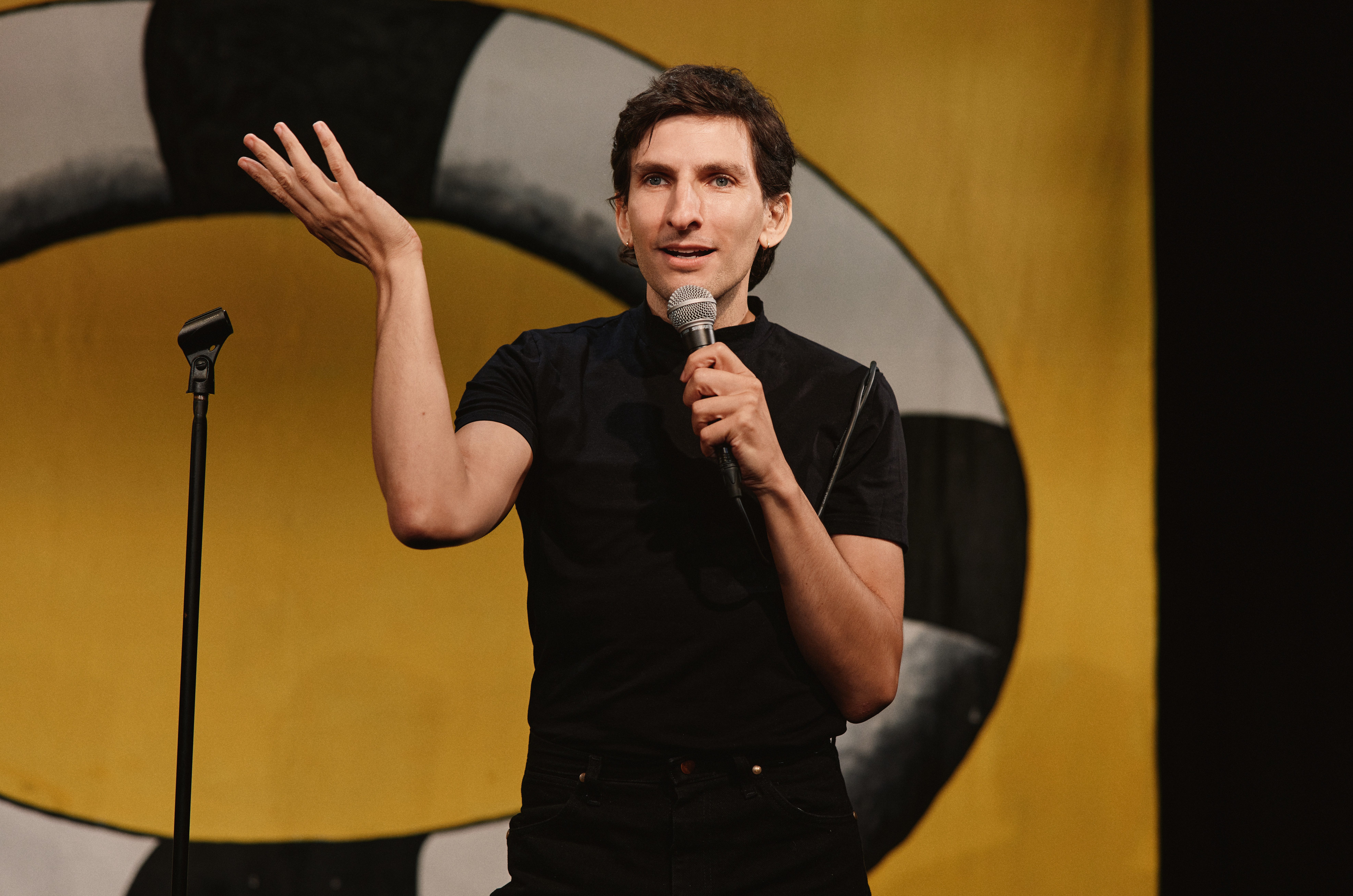 A man in all black standing onstage with a microphone in front of a painting of a life preserver