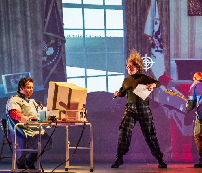 A man at a computer and a woman jumping on a colorful set 