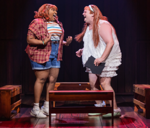 A Black woman and a white man in dresses wearing bright red wigs and laughing