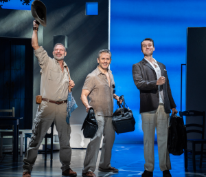Three middle-aged men standing and smiling with one waving his hat in the air
