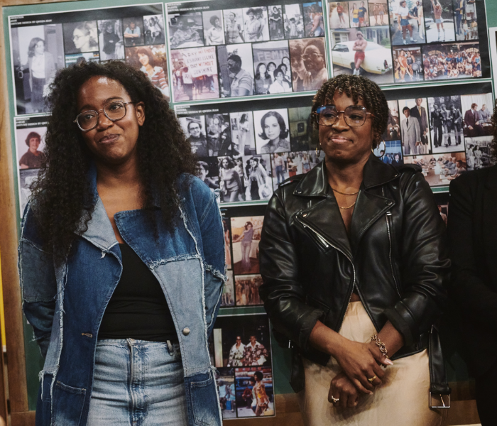 Whitney White and Kristolyn Lloyd standing next to each other smiling in front of a wall of photographs