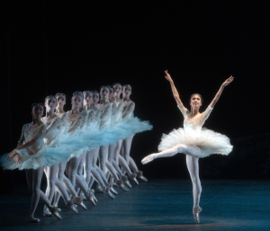A ballerina in a white leotard and tutu on pointe with her arms aloft with a row of ballerinas in blue on the side