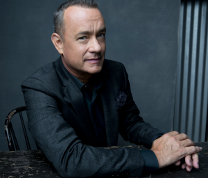 A photo of Tom Hanks leaning on a desk and looking at the camera with a slight smile against a gray background