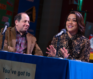 A man and a woman sitting at a desk smiling and talking into a microphone