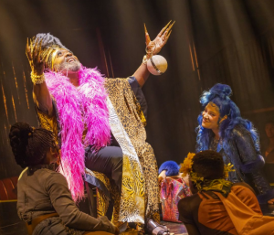 A performer draped in a pink boa extending his arms as other performers crouch befor him