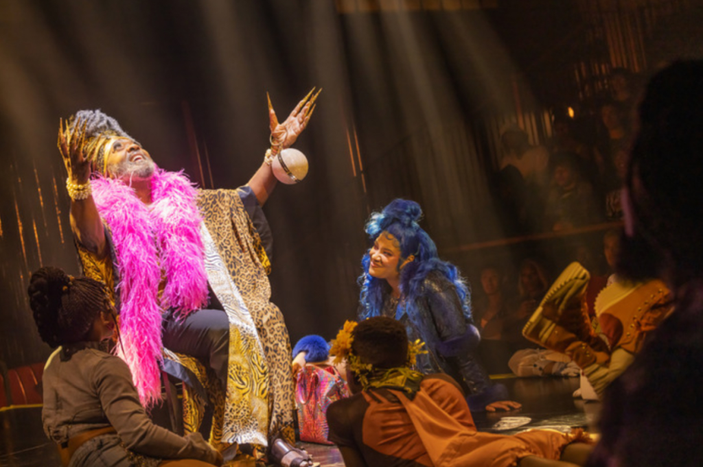 Junior LaBeija in a pink boa extending his arms surrounded by the ensemble 