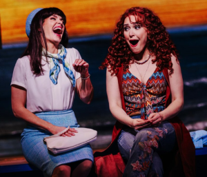 A woman in a blue hat and a woman with unruly red curls singing and smiling