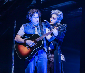 A young man playing an acoustic guitar as a black-clad man stands behind him