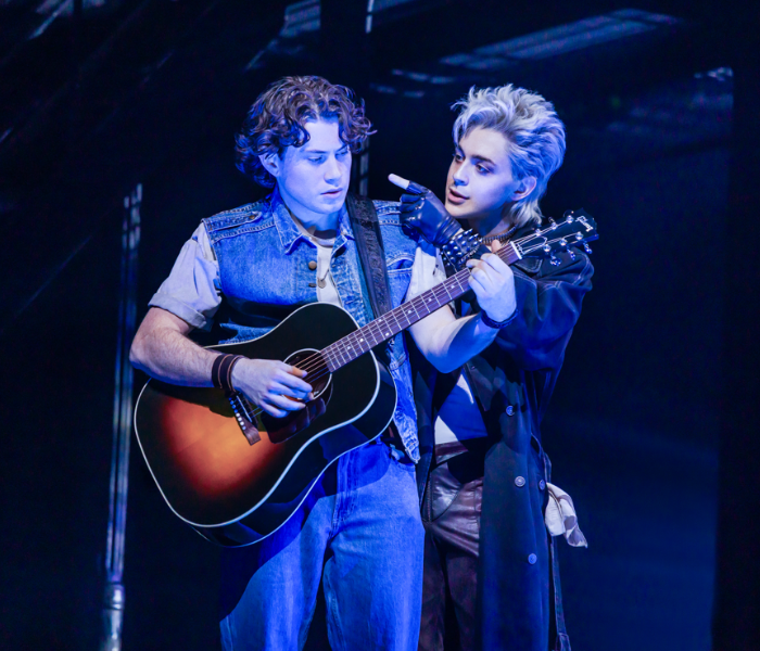 A young man playing an acoustic guitar as a black-clad man stands behind him