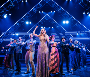 A performer playing Celine Dion pumping her fist in the air ith the cast of Titaníque behind her in blue light.