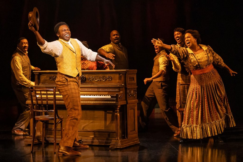 A man at a piano looking happy with others pointing at him