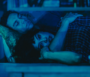 A man and a woman looking serious in blue light while lying in bed