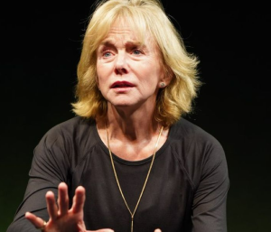 A close-up of a blonde woman looking serious and holding out her hand